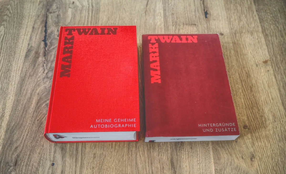 Zwei rote Bücher auf einer Holzoberfläche. Das linke Buch trägt den Titel „Meine Geheime Autobiographie“ von Mark Twain, und das rechte Buch trägt den Titel „Hintergründe und Zusätze“, ebenfalls von Mark Twain.