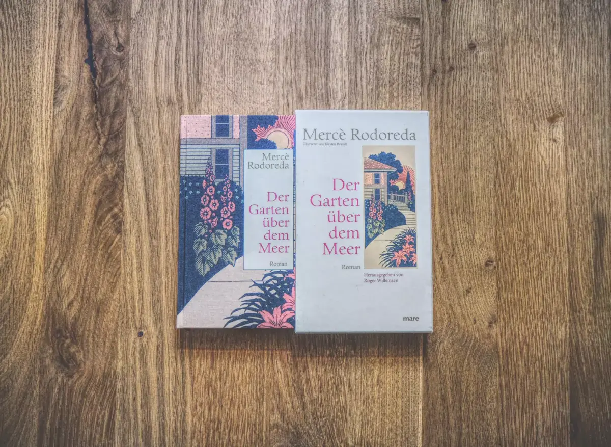 Zwei Exemplare von „Der Garten über dem Meer“ von Mercè Rodoreda, ein Taschenbuch und ein gebundenes Buch, nebeneinander auf einer Holzfläche platziert.