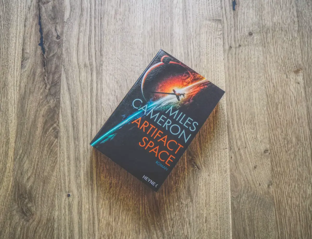 Ein Taschenbuch mit dem Titel „Artifact Space“ von Miles Cameron liegt auf einer Holzoberfläche.