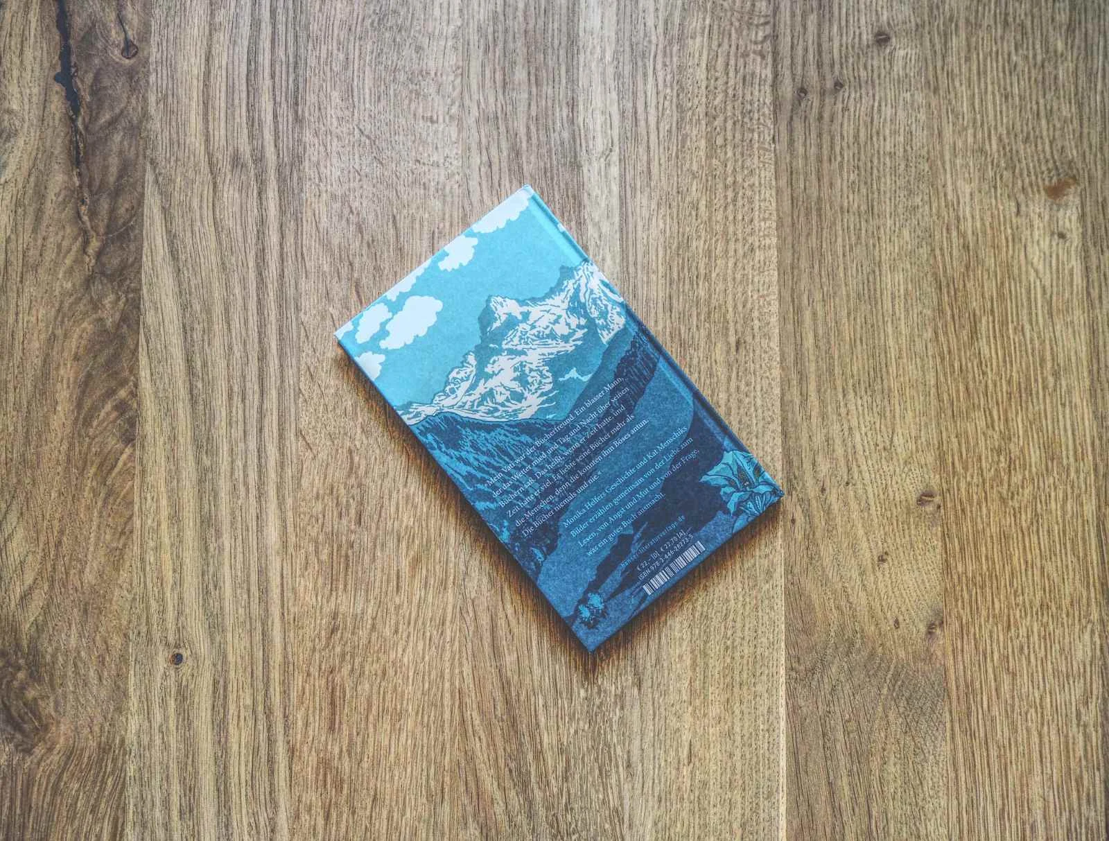 Ein geschlossenes Buch mit einer blau illustrierten Bergszene auf dem Einband liegt auf einer Holzfläche.