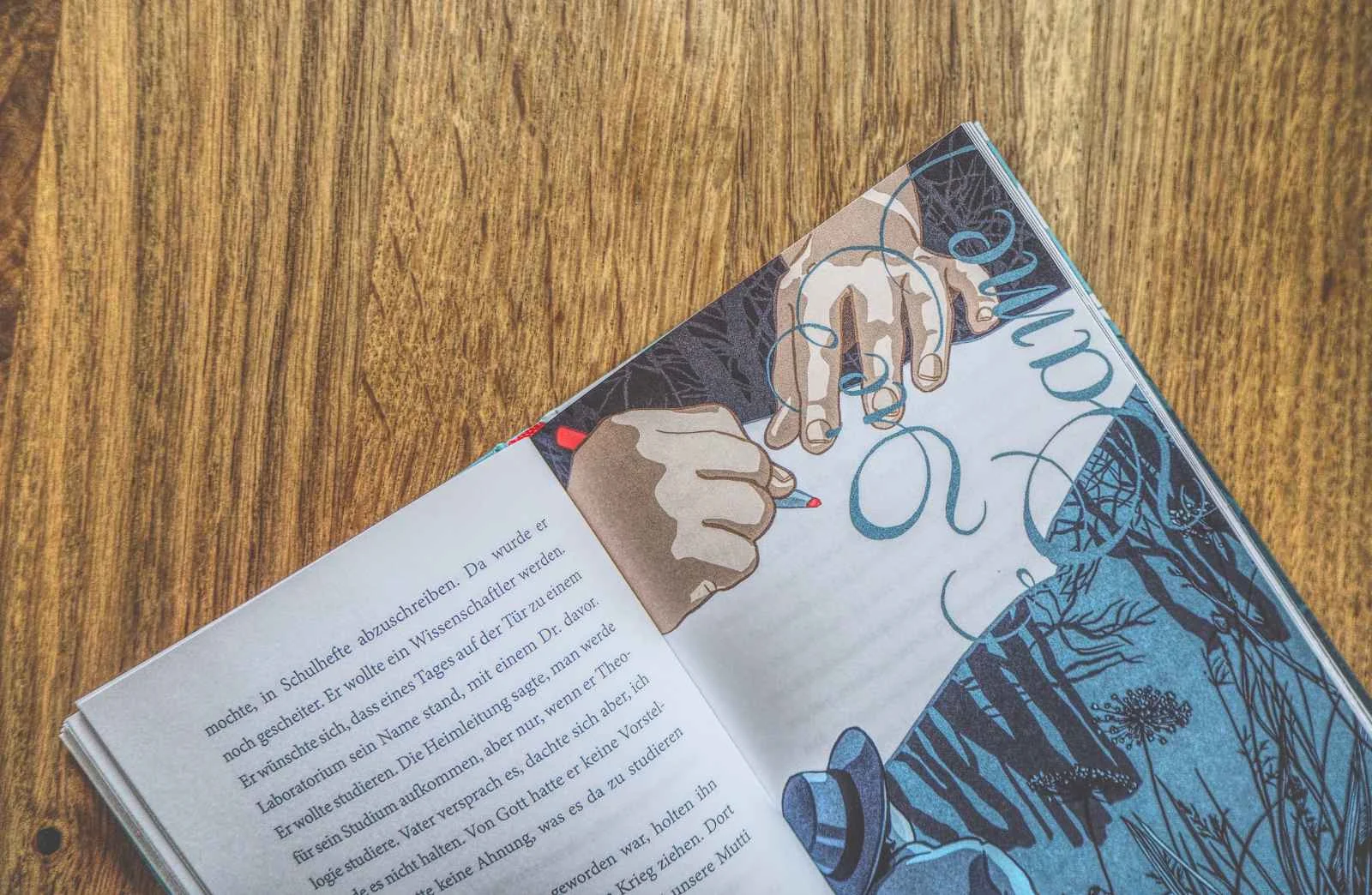 Ein auf einem Holztisch aufgeschlagenes Buch zeigt eine Illustration von zwei Händen, die mit roten Stiften zeichnen, sowie einen deutschen Text auf der gegenüberliegenden Seite.