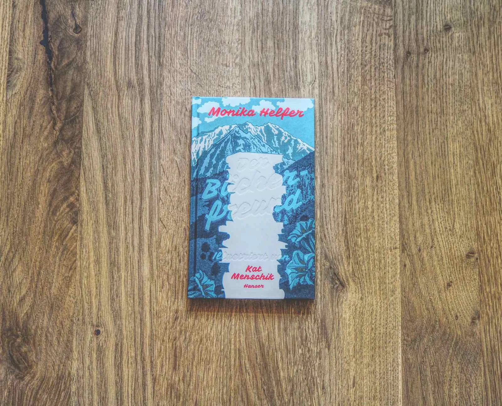 Ein Buch mit dem Titel "Der Bücherfreund" von Monika Helfer liegt auf einer Holzfläche. Der Einband zeigt blaue und weiße Illustrationen mit rotem und weißem Text.