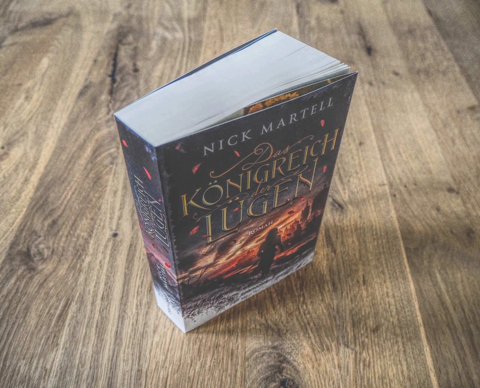 Ein Buch mit dem Titel „Das Königreich der Lügen“ von Nick Martell liegt auf einer Holzoberfläche. Das Cover zeigt eine dunkle, dramatische Szene mit einer Silhouette vor einem feurigen Hintergrund.