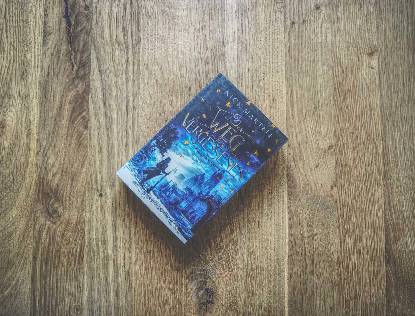 Auf einer Holzoberfläche liegt ein Buch mit dem Titel „Der Weg der Vergessenen“ von Nick Martell, dessen blau getönter Einband ein Mädchen und eine unheimliche Stadtlandschaft zeigt.