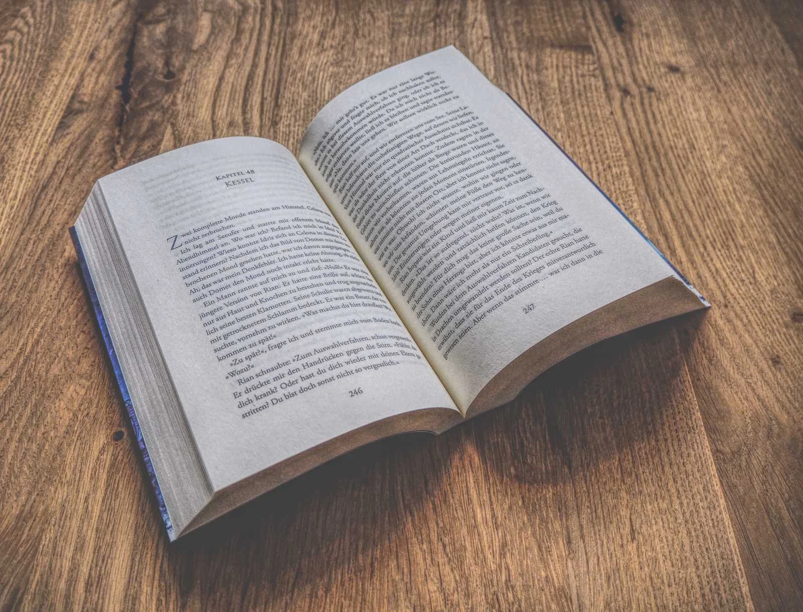 Auf einer Holzfläche liegt ein aufgeschlagenes Buch mit zweispaltigem Text.