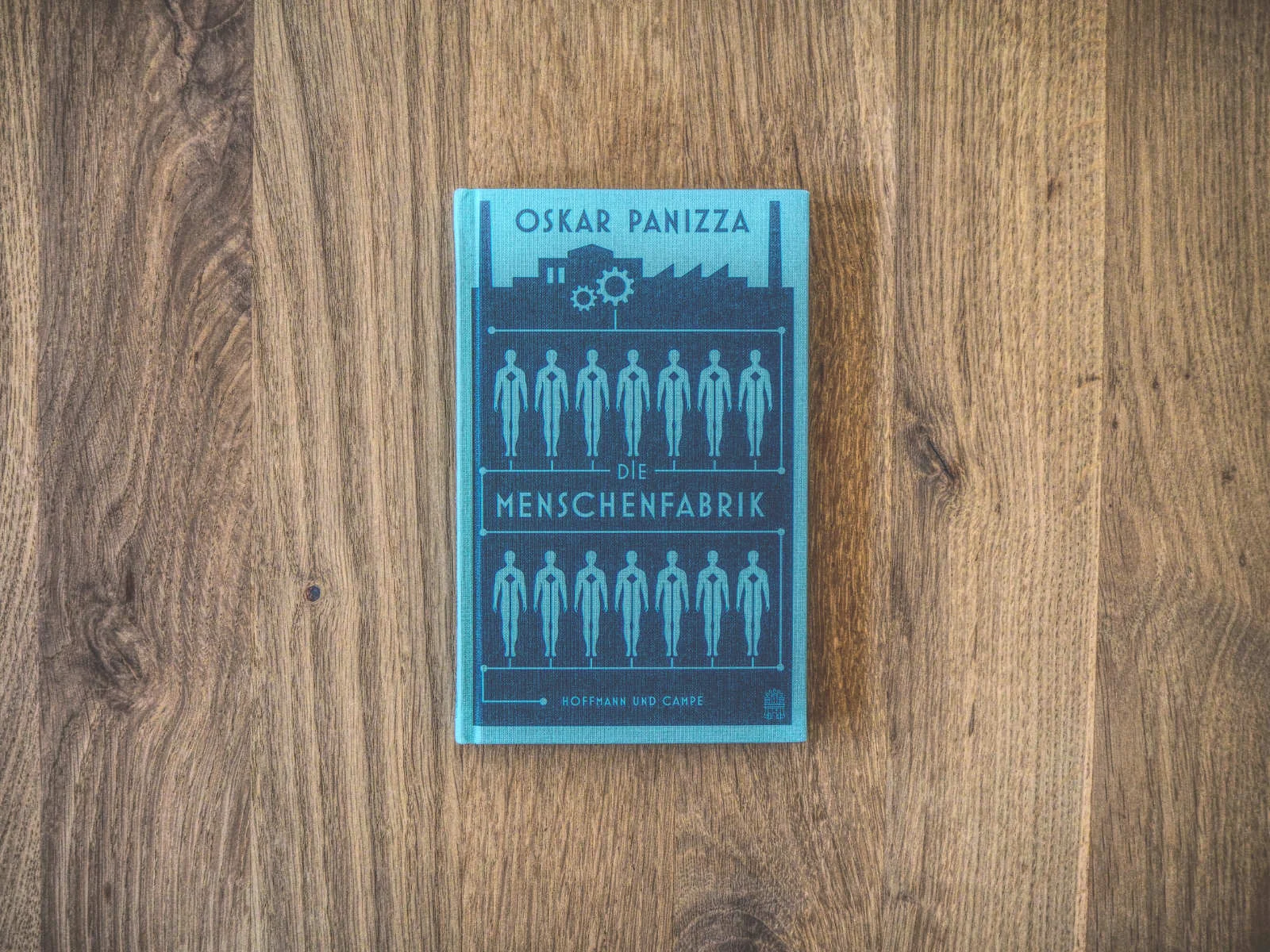 Auf einer Holzoberfläche liegt ein blaugrünes Buch mit dem Titel „Die Menschenfabrik“ von Oskar Panizza. Auf dem Cover ist eine Reihe menschlicher Figuren in einer industriellen Umgebung abgebildet.