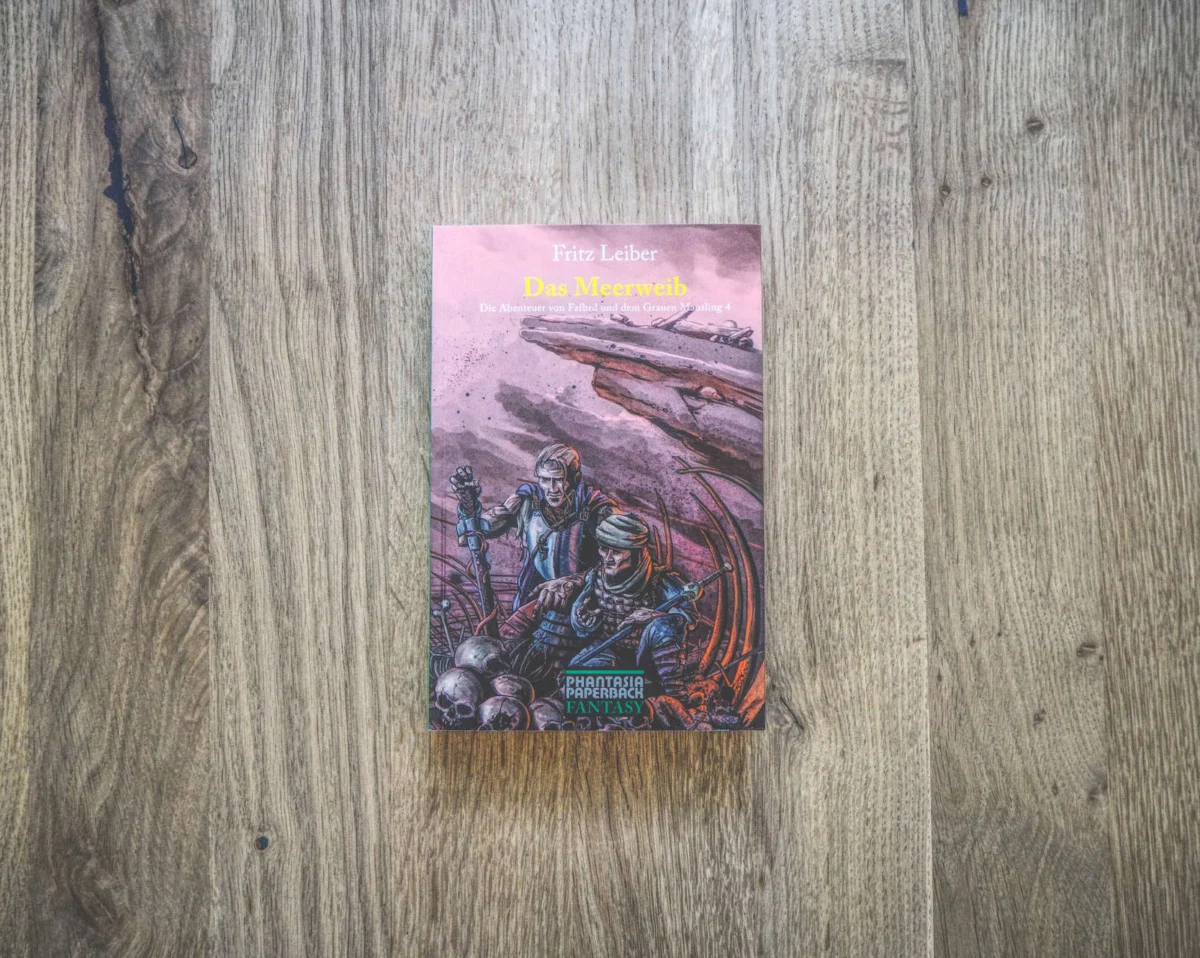 Ein Buch mit dem Titel „Fritz Leiber – Das Meeresthriller“ mit einer Fantasy-Coverillustration, die zwei Figuren zeigt, die zwischen Totenköpfen und Knochen stehen, die auf einer Holzoberfläche platziert sind.