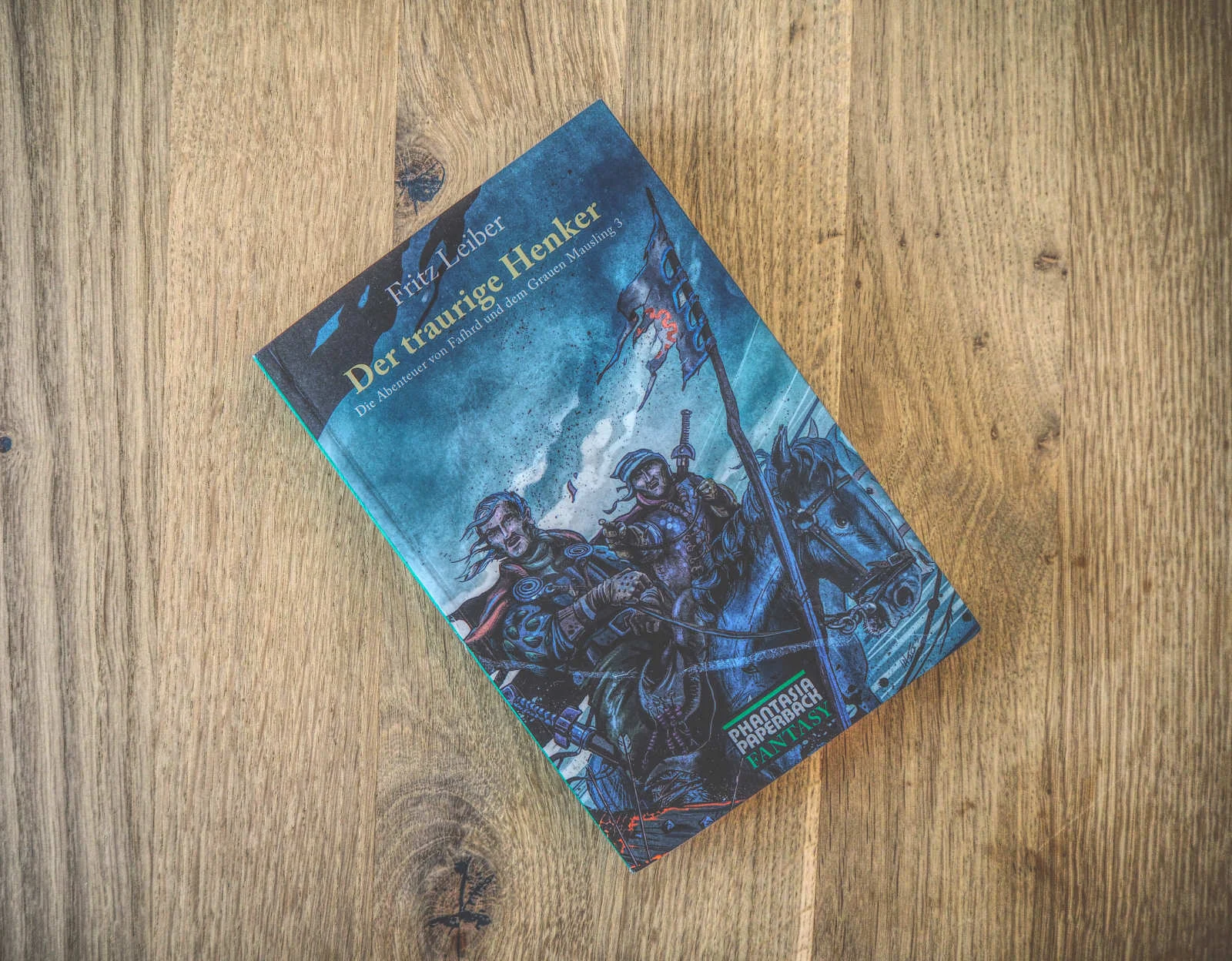 Ein Buch mit dem Titel „Der traurige Henker“ von Fritz Leiber liegt auf einer Holzfläche. Auf dem Cover sind zwei Soldaten in einer düsteren Kriegskulisse abgebildet.