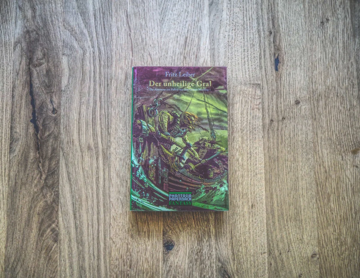 Auf einer Holzplatte liegt das Buch „Der unheilige Gral“ von Fritz Leiber. Auf dem Cover sind Figuren in einer dynamischen Fantasy-Szene abgebildet.