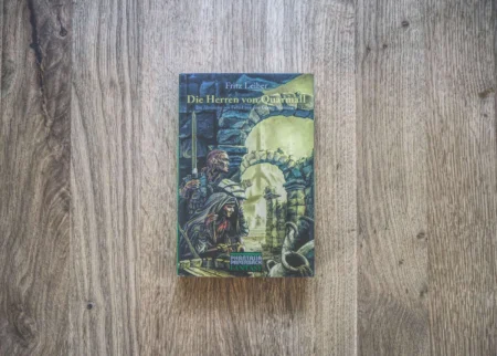 Ein Buch mit dem Titel „Die Herren von Quarmall“ von Fritz Leiber liegt auf einer Holzfläche. Das Cover zeigt eine illustrierte Fantasyszene mit Figuren in mittelalterlicher Kulisse.