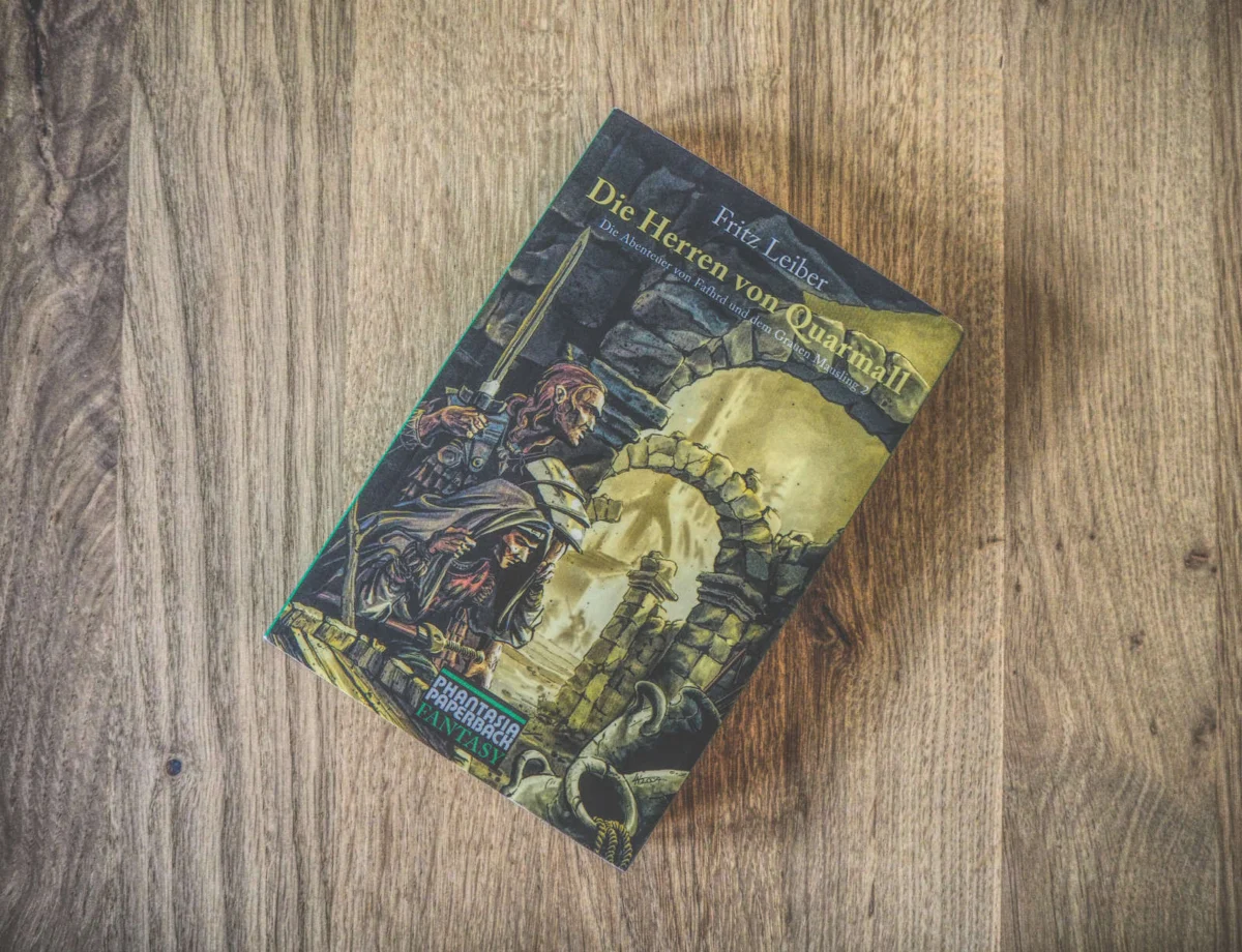 Ein Buch mit dem Titel „Die Herren von Quarmall“ von Fritz Leiber mit einem illustrierten Cover, das einen Krieger in Rüstung und eine Steinstruktur zeigt, die auf einer Holzoberfläche platziert sind.