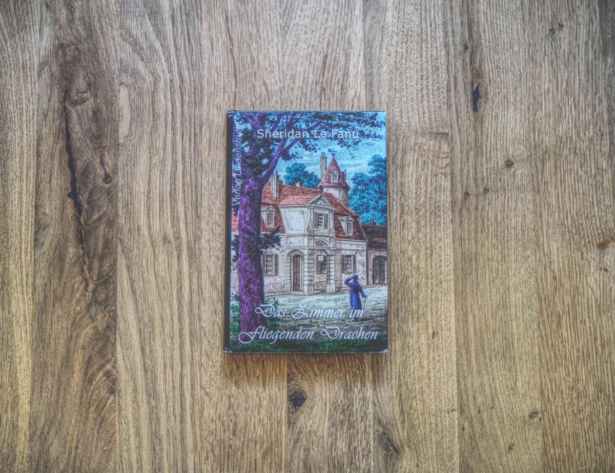 Auf einer Holzfläche liegt ein gebundenes Buch. Auf dem Cover ist ein altes Haus abgebildet, davor ein Mann. Der Titel lautet „Das Zimmer im fliegenden Drachen“ von Sheridan Le Fanu.