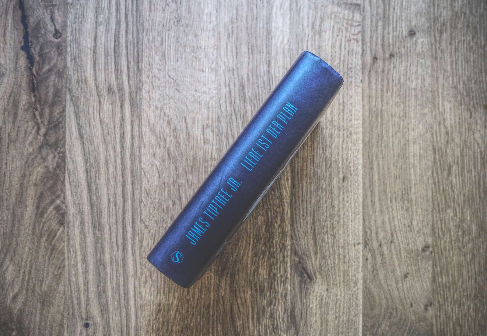 Ein Buch mit marineblauem Buchrücken, auf dem der Name des Autors, James Tiptree Jr., und der Titel „Liebe ist der Plan“ stehen, liegt auf einer Holzoberfläche.