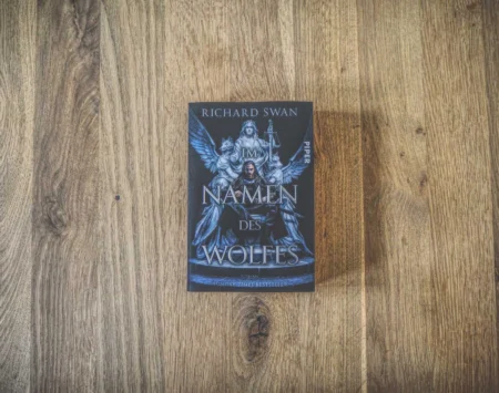 Eine deutsche Ausgabe des Buches „Namen des Wolfes“ von Richard Swan, auf dem Cover ist ein Krieger mit einer Axt abgebildet, der auf einer Holzoberfläche platziert ist.