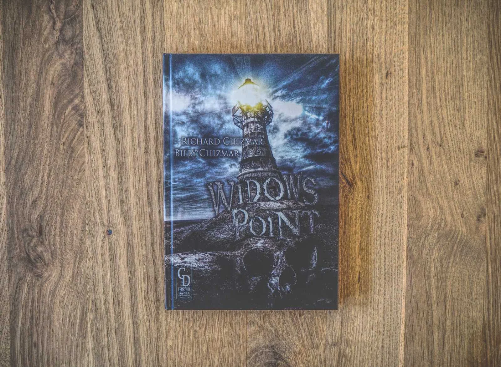 Das Bild zeigt das Buch „Widow's Point“ von Richard Chizmar und Billy Chizmar auf einer Holzoberfläche. Das Cover zeigt einen leuchtenden Leuchtturm an einer felsigen Küste unter einem stürmischen Himmel.