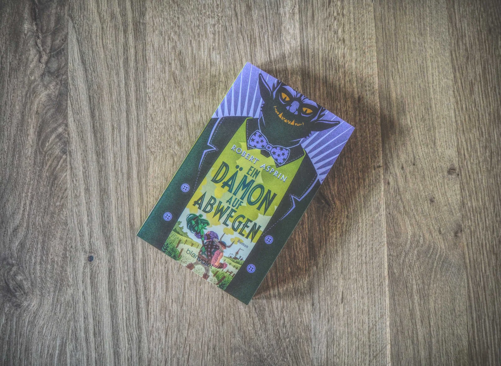 Auf einer Holzfläche liegt das Buch „Ein Dämon auf Abwegen“ von Robert Asprin. Auf dem Cover ist ein grinsender Dämon im Anzug abgebildet.