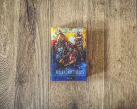Auf einer Holzfläche liegt ein gebundenes Buch mit dem Titel „Der Löwe von Tiberias“ von Robert E. Howard. Auf dem Cover ist ein Ritter auf einem Pferd abgebildet.