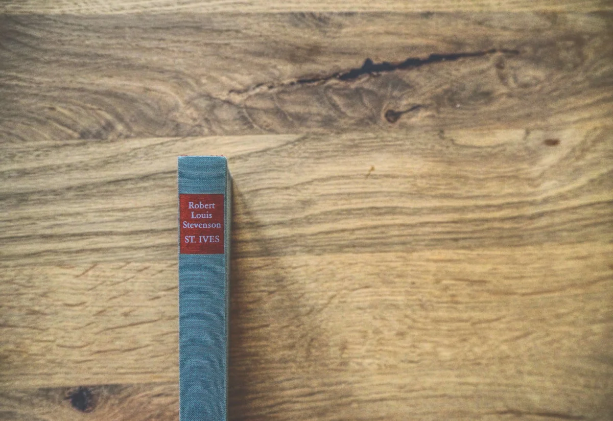 Ein Buch mit dem Titel „St. Ives“ von Robert Louis Stevenson, mit blauem Einband und rotem Rücken, steht aufrecht auf einer Holzfläche.
