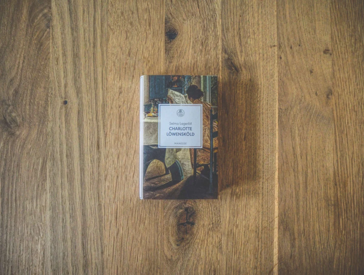 Auf einer Holzfläche liegt ein gebundenes Buch mit dem Titel „Charlotte Löwensköld“ von Selma Lagerlöf. Auf dem Cover ist eine Person abgebildet, die an einem Tisch sitzt und eine Zeitung liest.