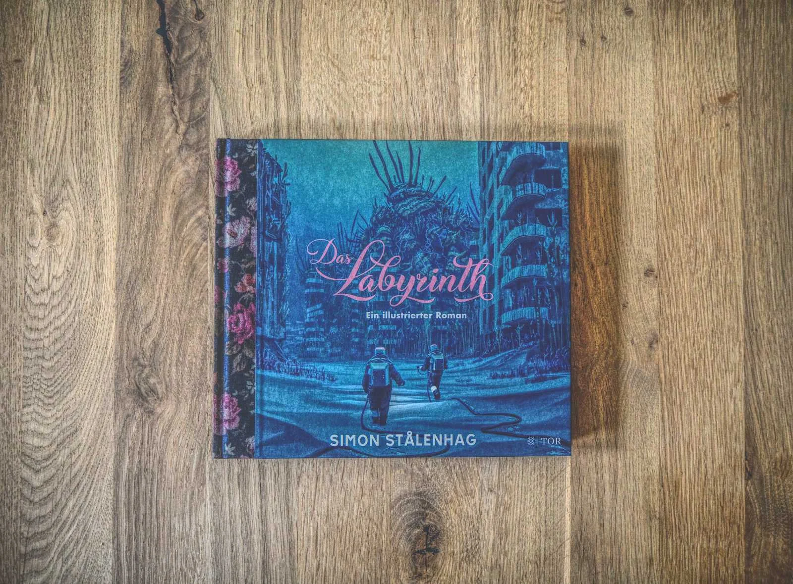 Ein Buch mit dem Titel „Das Labyrinth“ von Simon Stålenhag liegt auf einer Holzoberfläche. Das Cover zeigt zwei Figuren, die vor einem blau getönten, dystopischen Hintergrund auf ein großes, bedrohliches Bauwerk zulaufen.