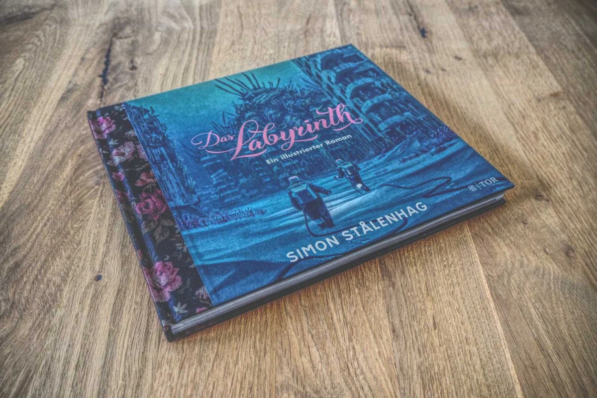 Auf einer Holzfläche liegt ein Buch mit dem Titel „Das Labyrinth“ von Simon Stålenhag. Auf dem Cover sind zwei Figuren zu sehen, die durch eine dystopische Landschaft wandeln.