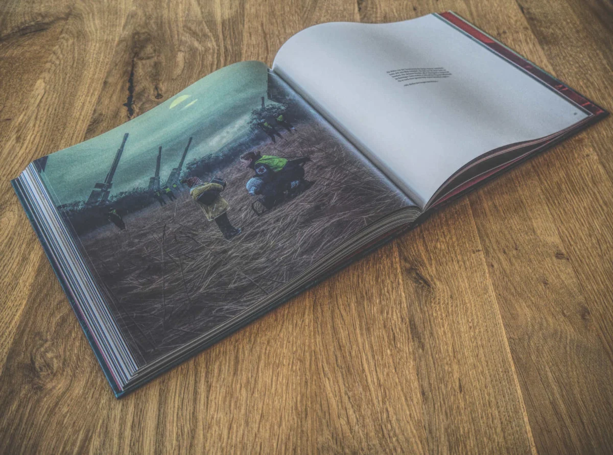 Ein offenes Buch auf einem Holztisch zeigt ein Foto von Menschen, die unter einem bewölkten Himmel auf einem Feld arbeiten, sowie eine Seite mit Text.