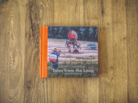 Auf einer Holzoberfläche liegt ein gebundenes Buch mit dem Titel „Tales from the Loop“ von Simon Stålenhag. Auf dem Cover ist ein Kind neben einem Roboter und einem Auto abgebildet.