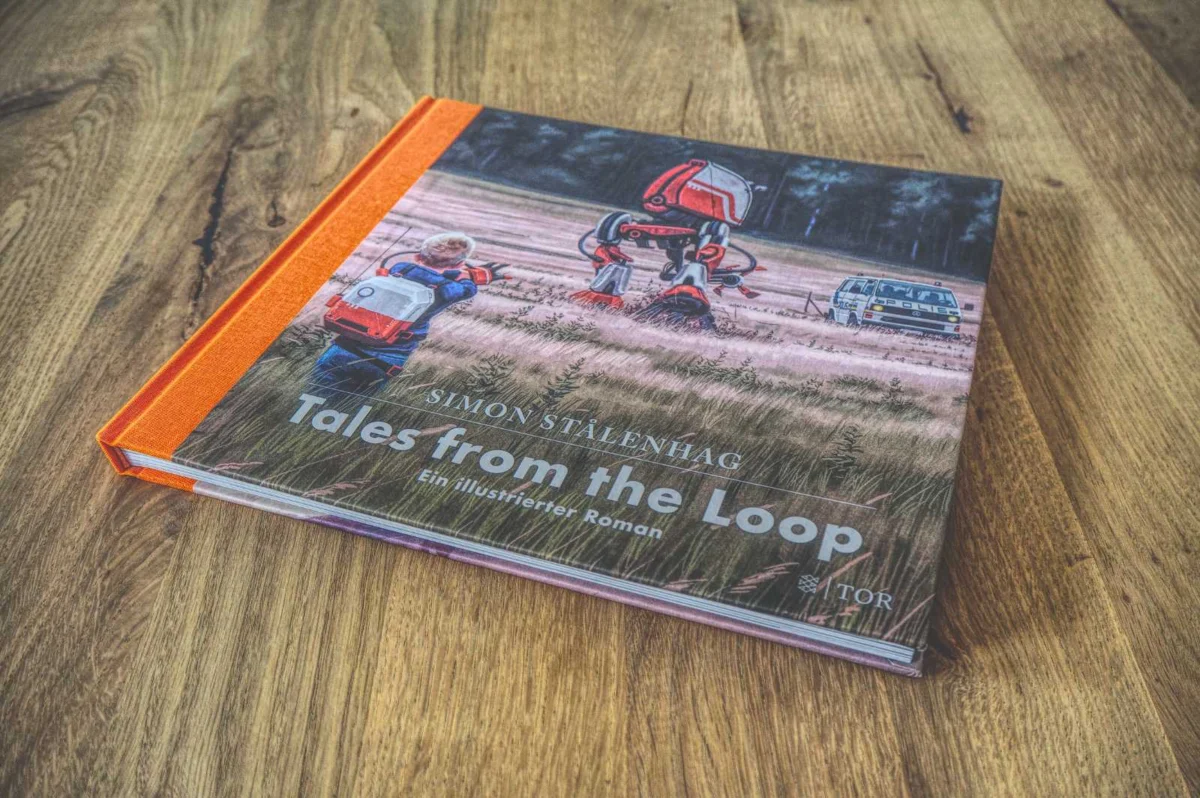 Nahaufnahme des Buches „Tales from the Loop“ von Simon Stalenhag auf einer Holzoberfläche. Das Cover zeigt Kinder und einen Roboter in einer ländlichen Landschaft.