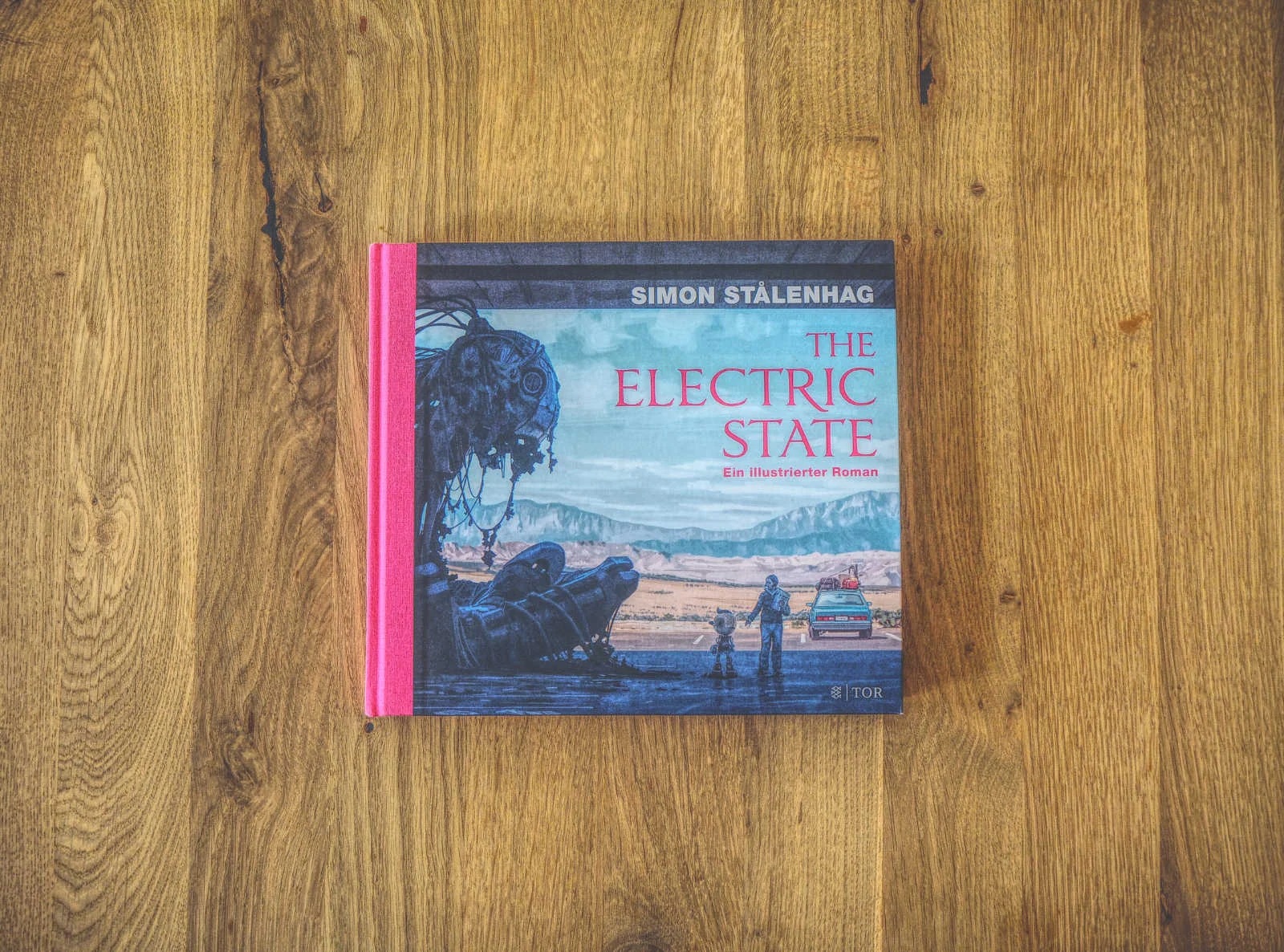 Auf einer Holzoberfläche liegt ein Buch mit dem Titel „The Electric State“ von Simon Stålenhag. Der Buchrücken ist rosa. Auf dem Cover ist ein beschädigter Roboter abgebildet, daneben steht eine Person.