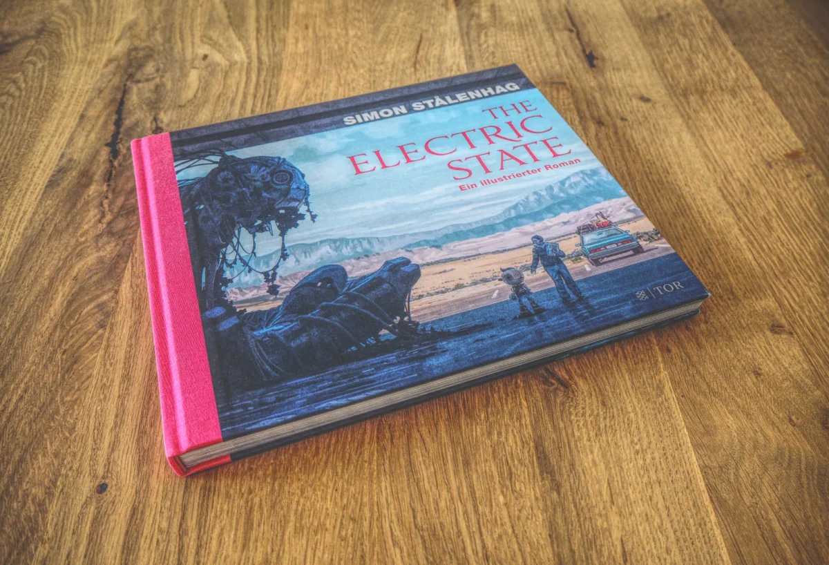 Ein gebundenes Buch mit dem Titel „The Electric State“ von Simon Stålenhag mit rotem Buchrücken, auf einer Holzoberfläche platziert.