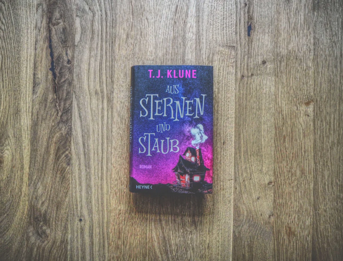 Auf einer Holzfläche liegt das deutschsprachige Buch „Aus Sternen und Staub“ von T.J. Klune. Auf dem Cover ist ein mystisches Haus mit Rauch vor einem sternenklaren Nachthimmel abgebildet.