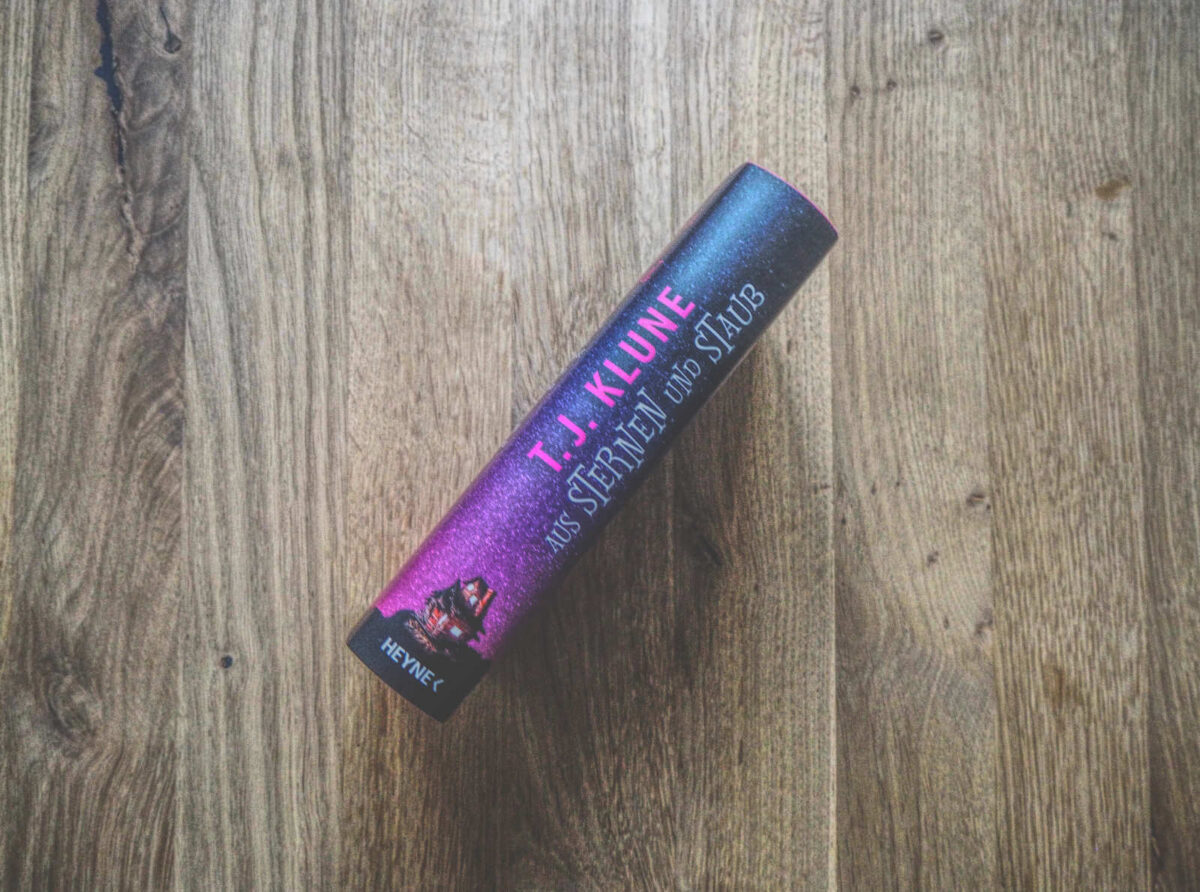 Ein Buch mit dem Titel „Aus Sternen und Staub“ von T. J. Klune liegt mit dem Buchrücken nach oben auf einer Holzfläche.