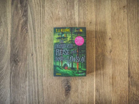 Das Cover des Buches „Die unerhörte Reise der Familie Lawson“ von T. J. Klune, auf einer Holzoberfläche. Das Cover zeigt eine Waldszene mit einem Haus und einer Familie.