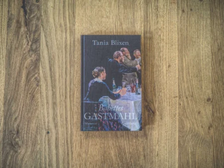 Ein Foto des Buches „Babettes Gastmahl“ von Tania Blixen, platziert auf einer Holzoberfläche. Das Cover zeigt eine Abbildung von Menschen, die an einem Tisch speisen.