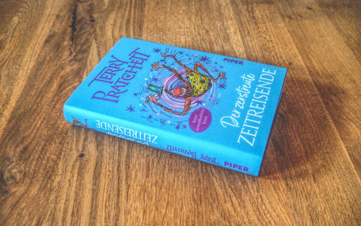 Auf einer Holzfläche liegt ein blaues Buch mit dem Titel „Der zerstreute Zeitreisende“ von Terry Pratchett.