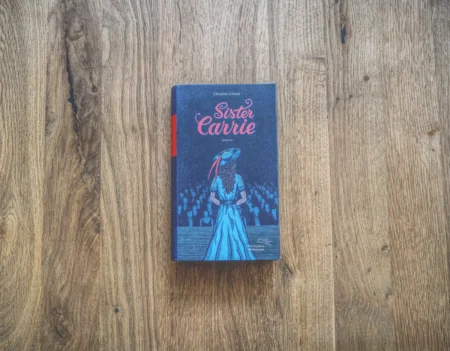 Ein Buch mit dem Titel „Sister Carrie“ von Theodore Dreiser mit einem blauen Einband und der Abbildung einer Frau mit Hut und Kleid, die vor einem Publikum steht, auf einer Holzoberfläche platziert.