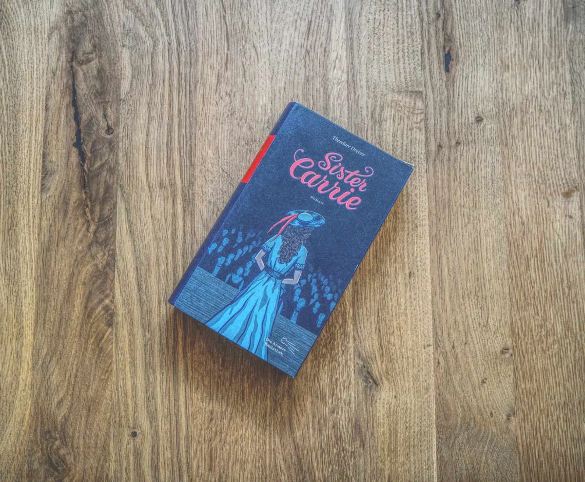 Ein gebundenes Buch mit dem Titel „Sister Carrie“ liegt auf einer Holzoberfläche. Die Coverillustration zeigt eine Frau mit Hut und Kleid vor einem dunklen Hintergrund.