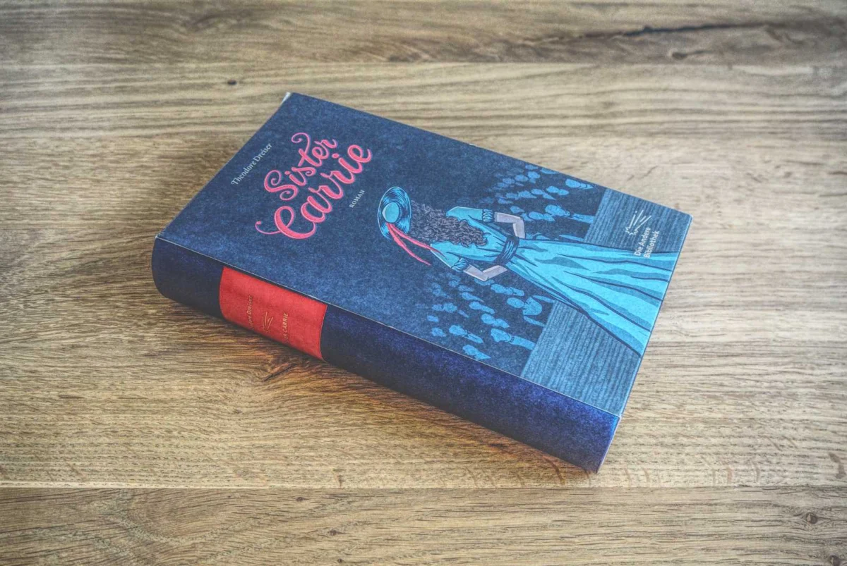 Auf einer Holzoberfläche liegt ein gebundenes Exemplar von „Sister Carrie“ von Theodore Dreiser. Auf dem Cover ist eine Frau in einem blauen Kleid mit einer roten Schleife im Haar abgebildet.