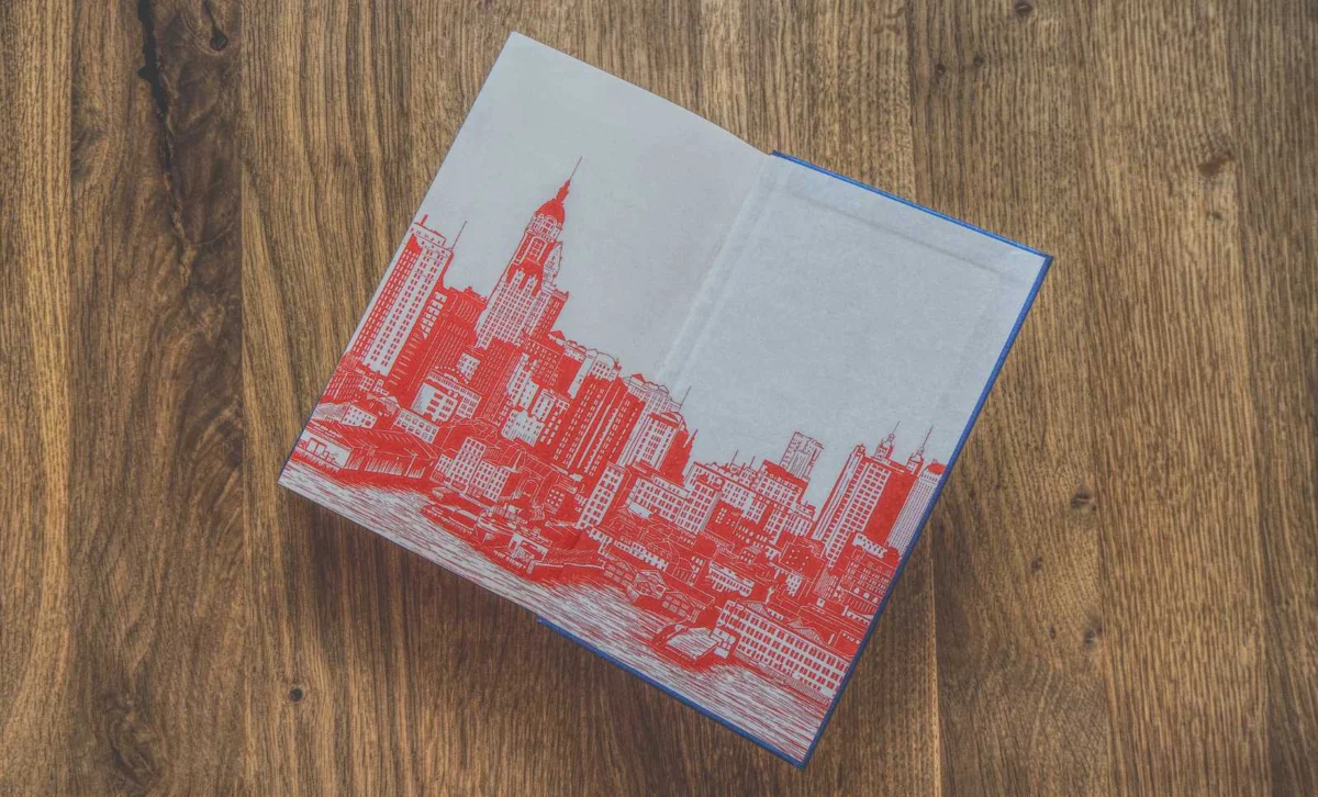 Auf einer Holzoberfläche liegt ein offenes Buch, das die überwiegend in roter Tinte gehaltene Illustration einer Stadtskyline zeigt.