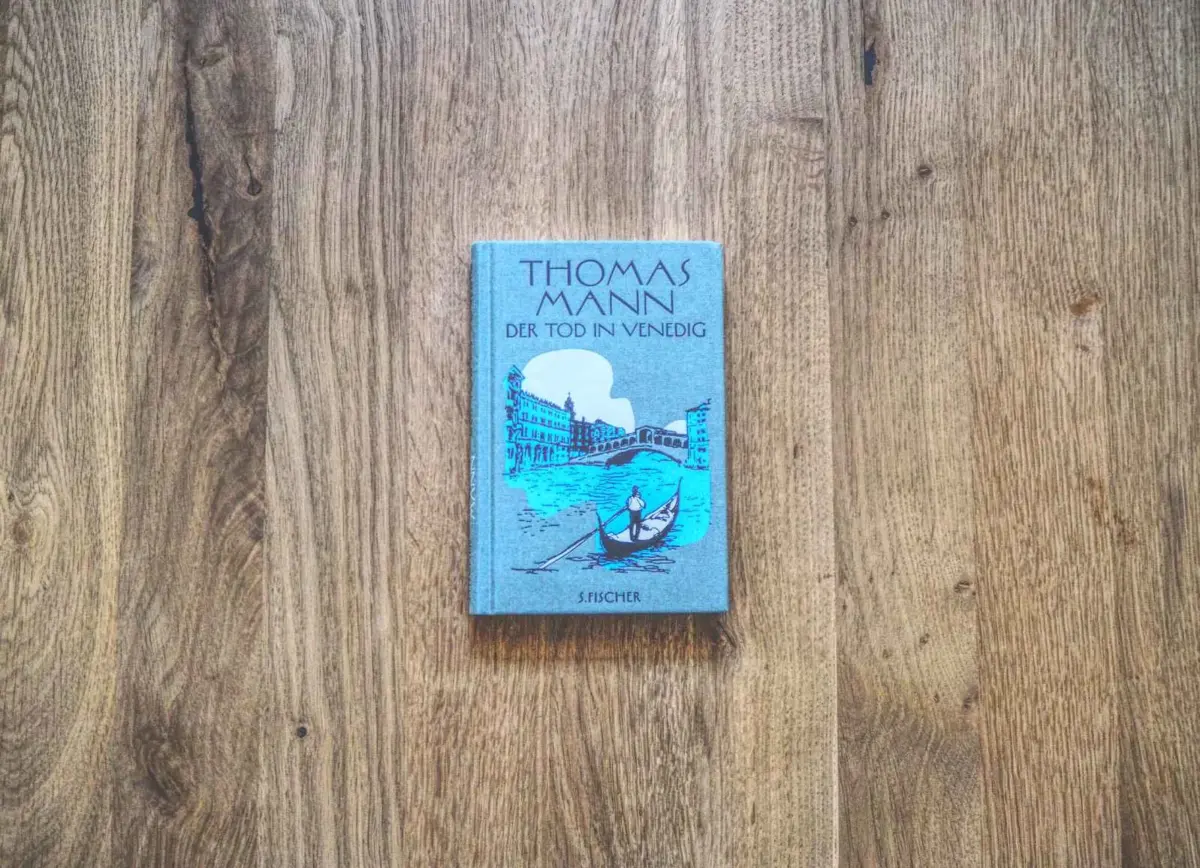 Das Bild zeigt das Buch „Der Tod in Venedig“ von Thomas Mann, platziert auf einer Holzplatte. Das Cover zeigt eine Abbildung von Venedig mit Gondeln.