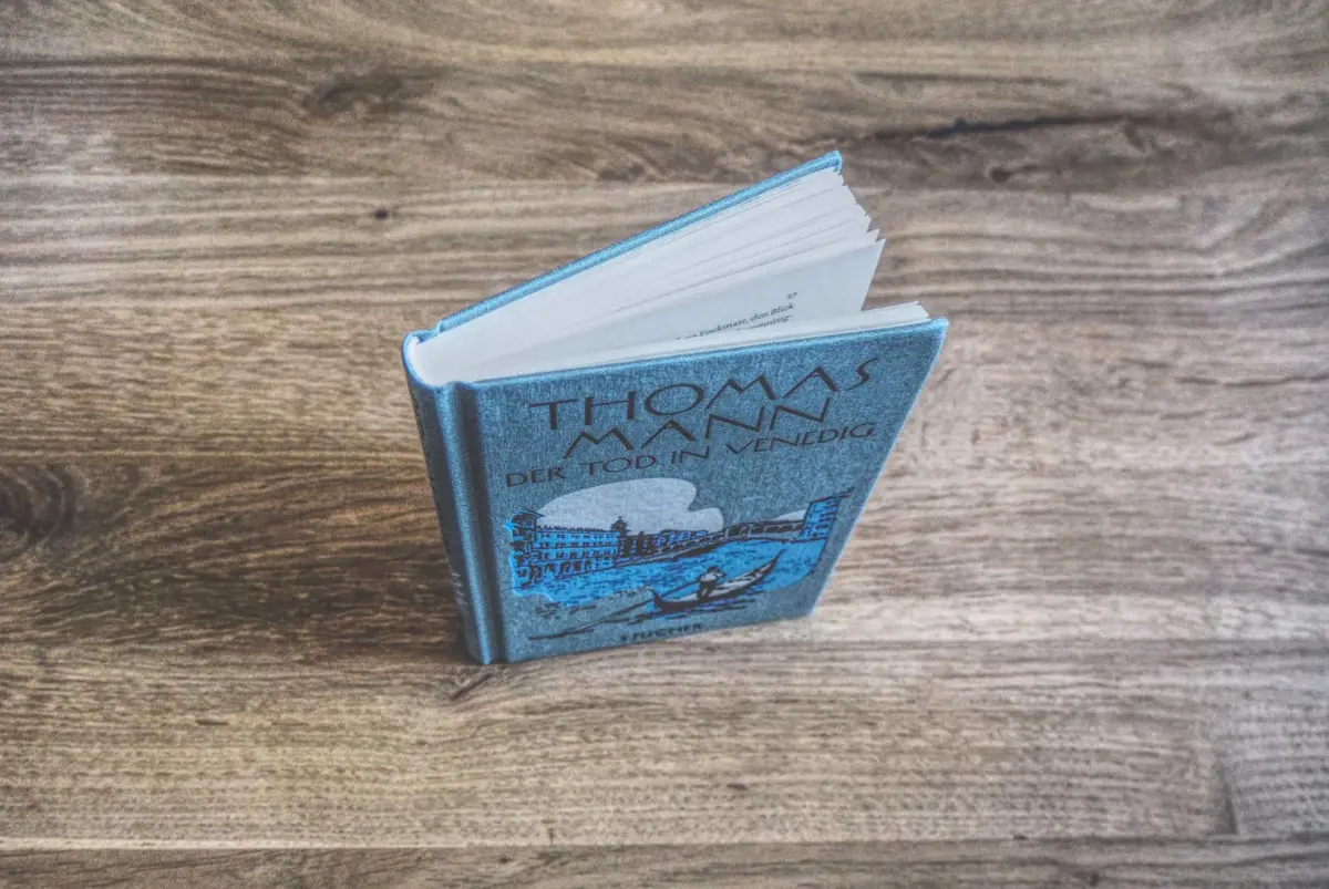Auf einer Holzplatte steht ein blau eingebundenes Buch mit dem Titel „Der Tod in Venedig“ von Thomas Mann. Auf dem Cover ist eine Venedig-Illustration zu sehen.