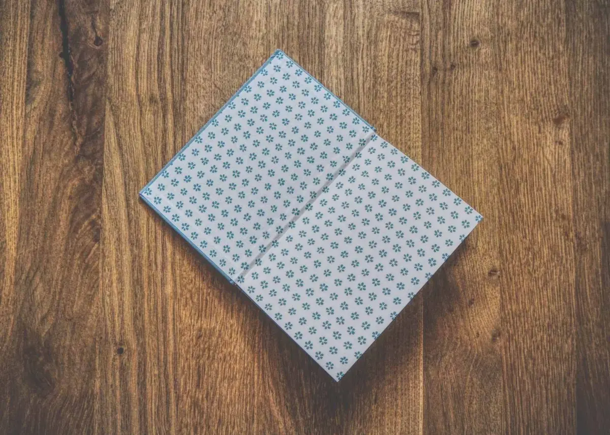 Auf einer Holzoberfläche liegt ein offenes Buch mit einem Einband mit blauem Blumenmuster.