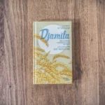 Das Bild zeigt das Buch „Djamila“ von Tschingis Aitmatow mit Illustrationen von Kat Menschik, platziert auf einer Holzoberfläche. Der Umschlag zeigt ein Muster aus gelben Weizenhalmen.