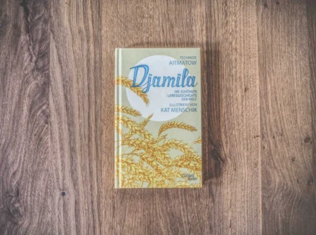 Das Bild zeigt das Buch „Djamila“ von Tschingis Aitmatow mit Illustrationen von Kat Menschik, platziert auf einer Holzoberfläche. Der Umschlag zeigt ein Muster aus gelben Weizenhalmen.