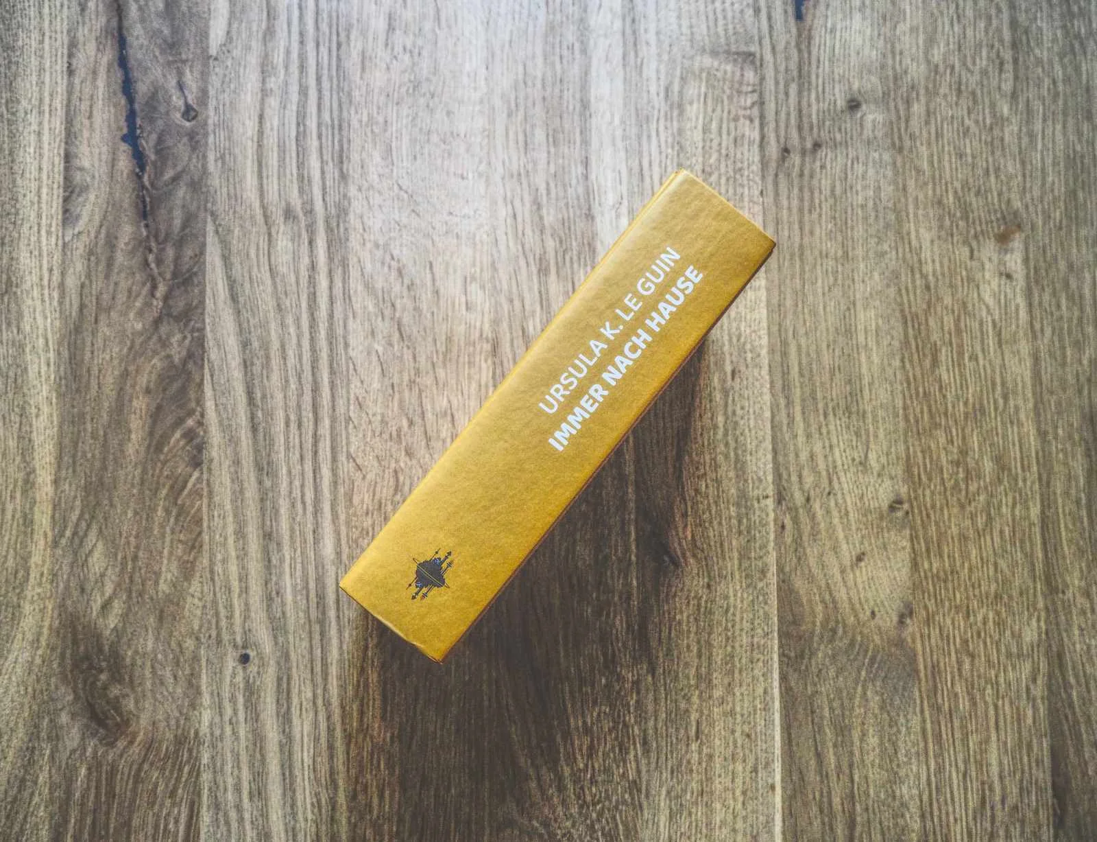 Ein gelbes, gebundenes Buch mit dem Titel "Immer nach Hause" von Ursula K. Le Guin liegt auf einer hölzernen Oberfläche, vom Buchrücken aus betrachtet.