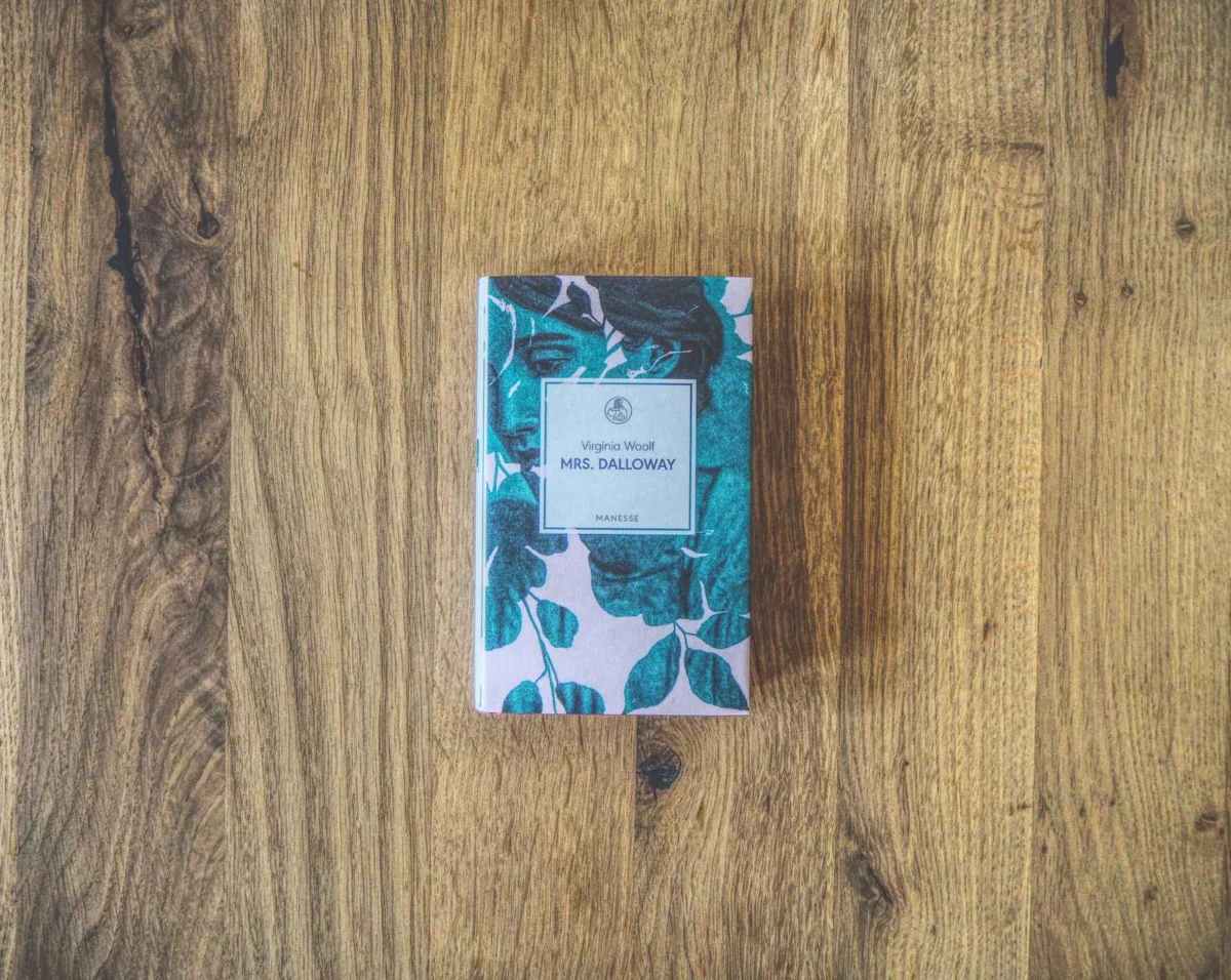 Auf einem Holztisch liegt ein gebundenes Exemplar von „Mrs. Dalloway“ von Virginia Woolf, eingebunden in einen Umschlag mit Blumenmuster.