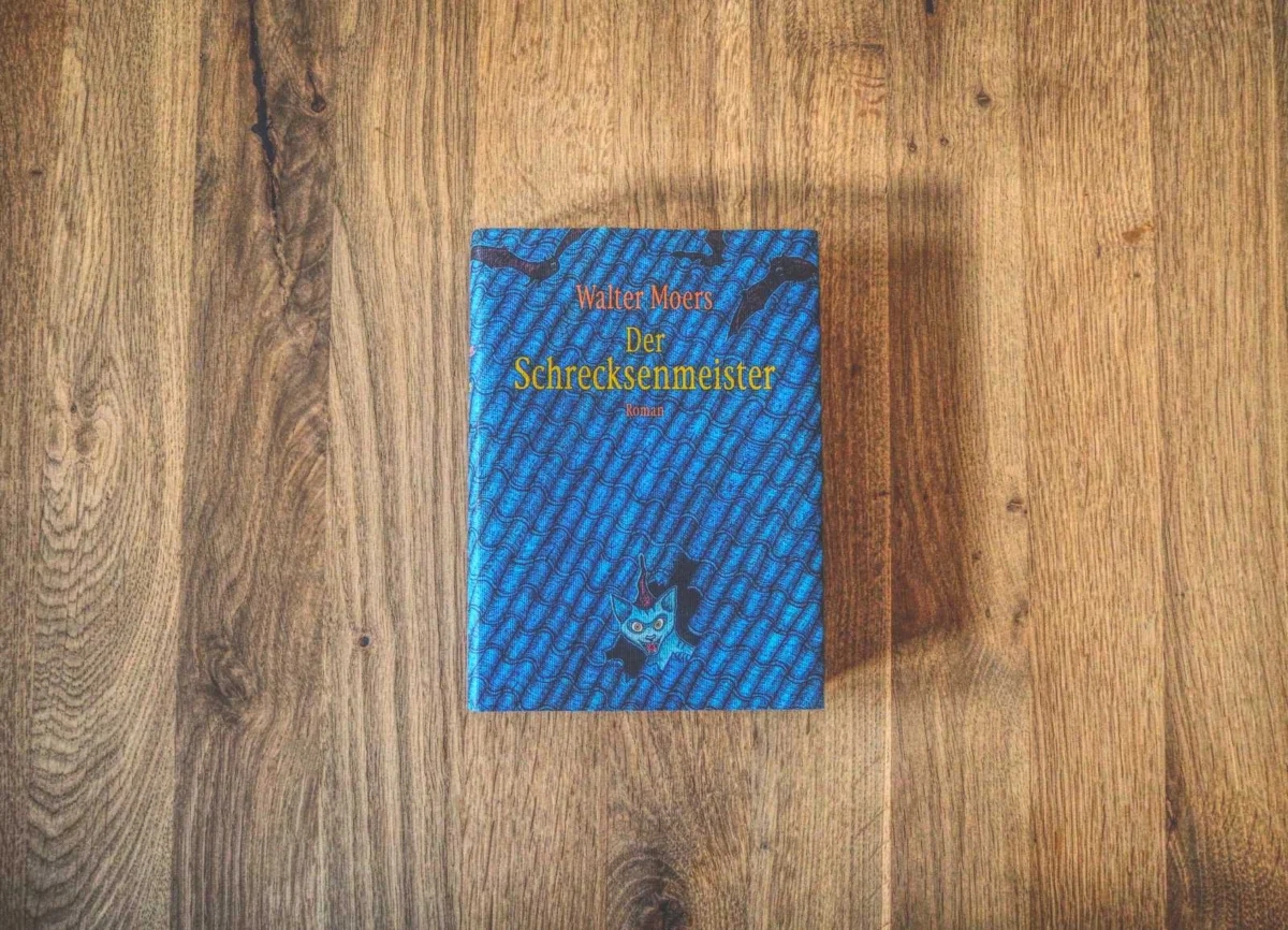 Ein Buch mit dem Titel „Der Schrecksenmeister“ von Walter Moers mit einem blauen, strukturierten Cover, in dessen unteren Bereich ein kleines Wesen abgebildet ist, das auf einer Holzoberfläche fotografiert ist.