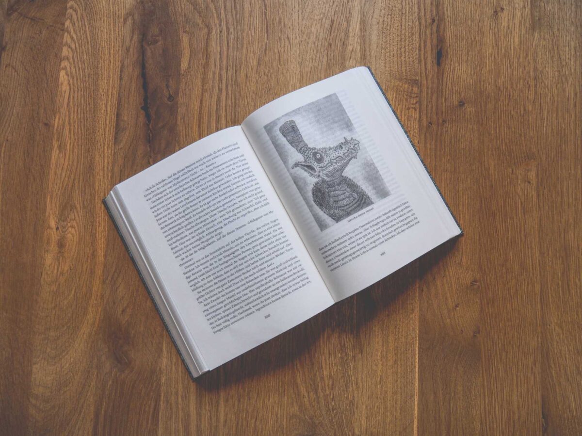 Ein aufgeschlagenes Buch auf einem Holztisch, auf dessen beiden Seiten Text zu sehen ist und auf der rechten Seite die Abbildung eines reptilienartigen Wesens mit einer Pfeife.