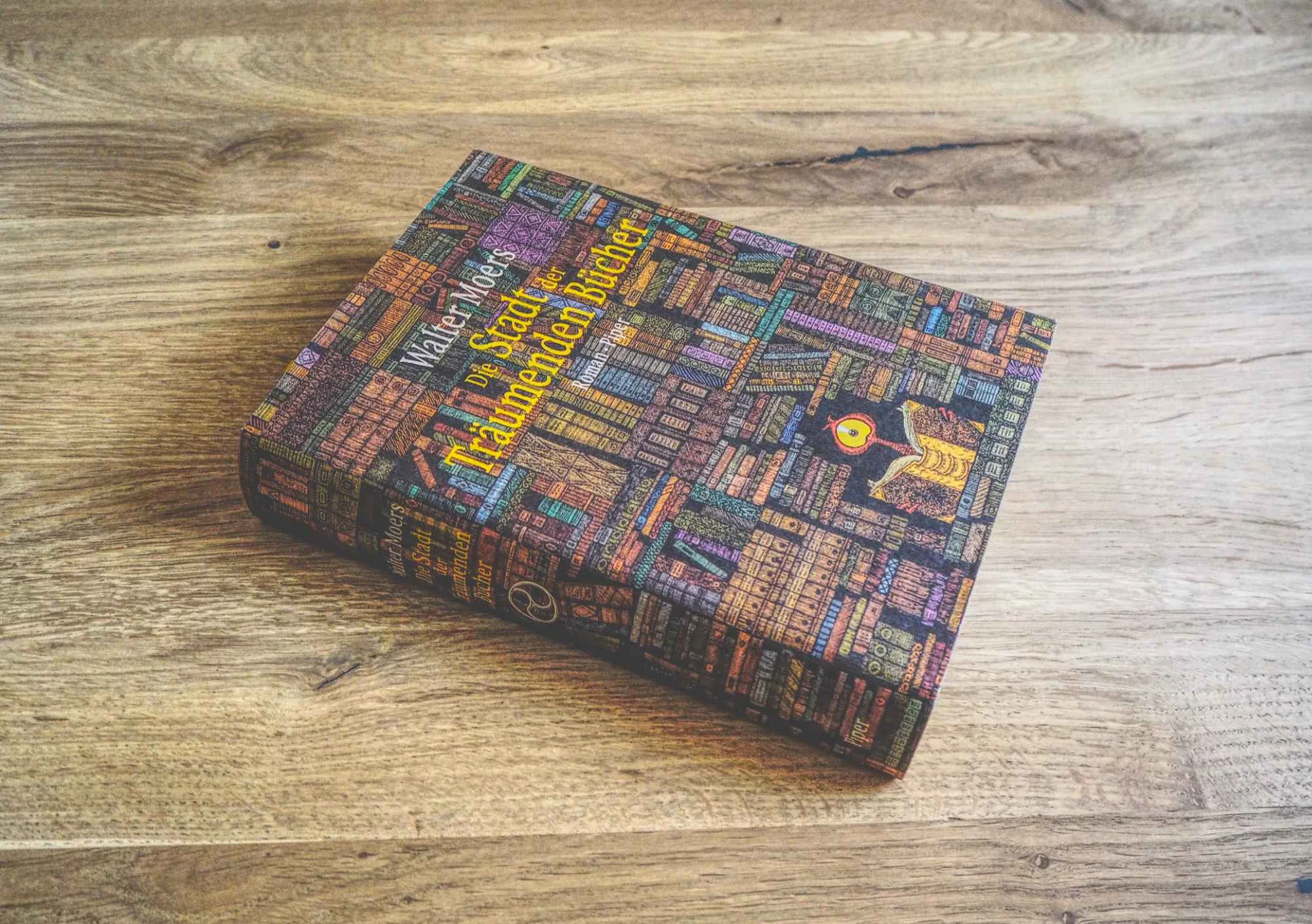 Ein gebundenes Buch mit dem Titel "Die Stadt der träumenden Bücher" von Walter Moers liegt auf einer Holzfläche. Das Cover zeigt eine Illustration von Bücherregalen, die mit bunten Büchern gefüllt sind.