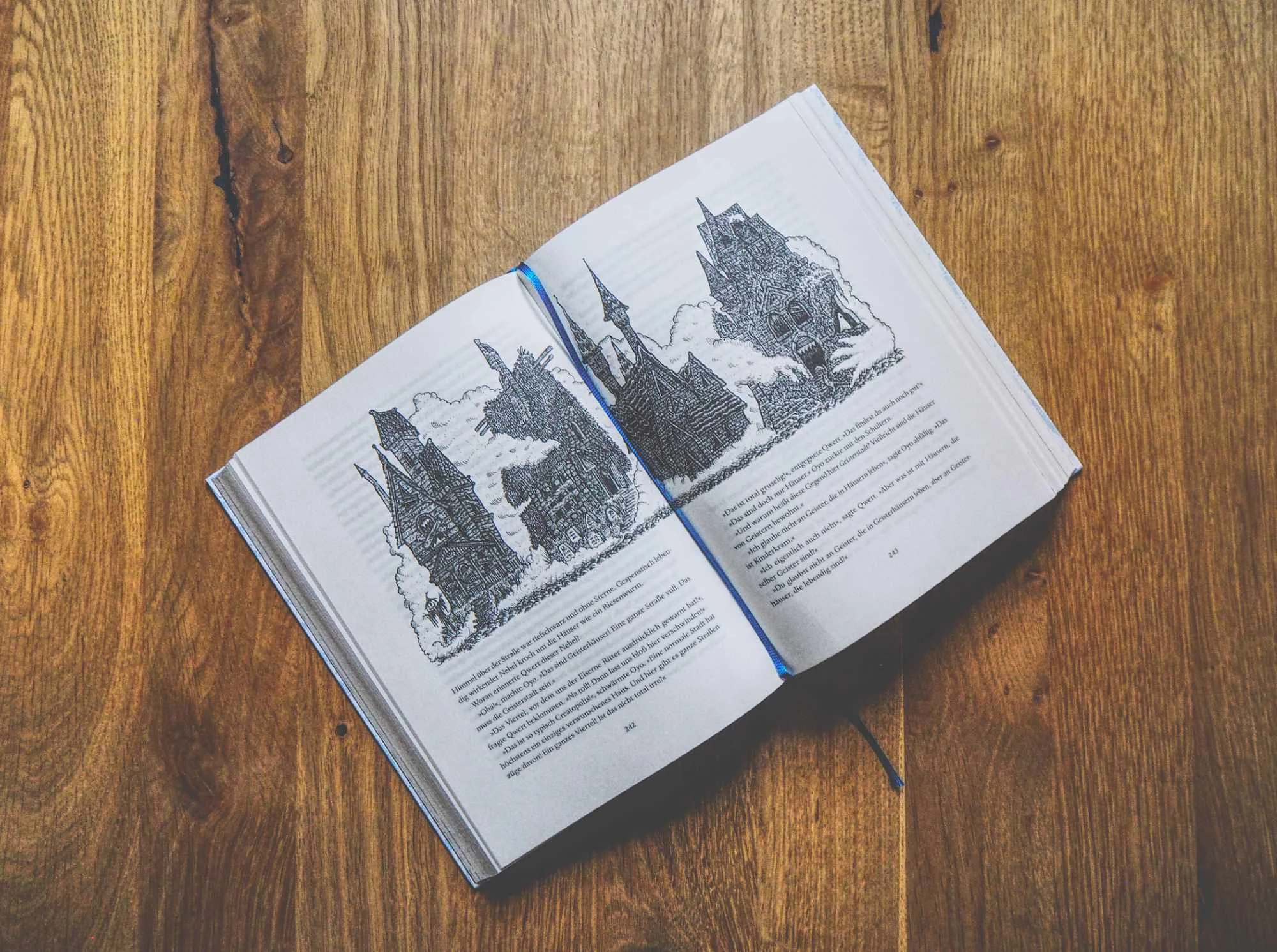 Ein aufgeschlagenes Buch mit Schwarz-Weiß-Abbildungen von Gebäuden liegt auf einer hölzernen Unterlage, in der Mitte befindet sich ein Lesezeichen mit blauem Band.